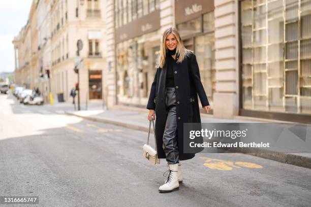 Xenia Adonts wears gold and rhinestones earring, a black wool turtleneck ribs open neck pullover, a long black Bottega Veneta coat, a white latte...