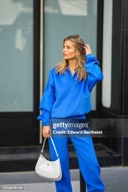 Xenia Adonts wears a gold earring, a royal blue V-neck sweater with matching large Shabderb jogger sport pants, a gold ring, a white shiny leather...