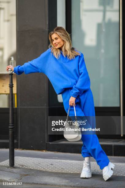 Xenia Adonts wears a pearls necklace, a royal blue V-neck sweater with matching large Shabderb jogger sport pants, a gold watch, a gold ring, a white...