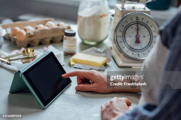 hand van een vrouw die een schort draagt die een levende kooksessie op haar tablet bekijkt en het recept volgt - baking reading recipe stockfoto's en -beelden