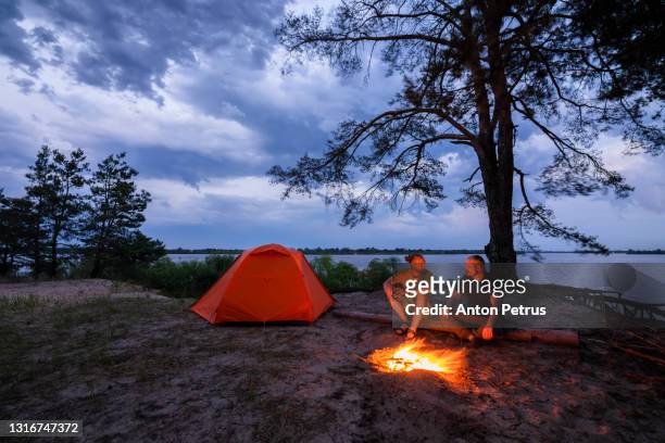 a couple of friends near the bonfire near the tent on the lake at sunset. weekend hike - bonfire beach stock pictures, royalty-free photos & images