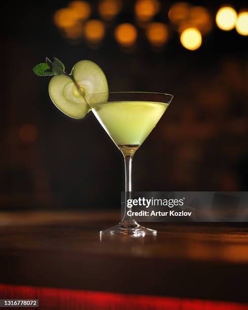 alcoholic drink cocktail with slice of green apple in martini glass. object at table on black background with beautiful bokeh. vertical format - green apple slices stock pictures, royalty-free photos & images