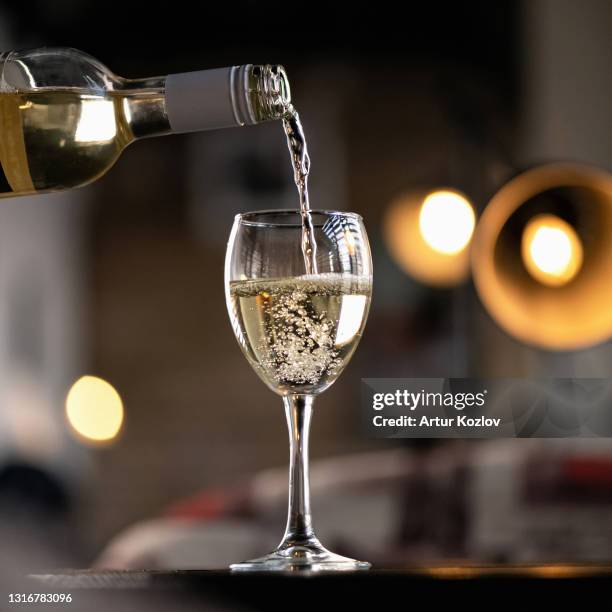 white wine pours from bottle into wine glass. alcoholic beverage with air bubbles on table against of backlit blurred background. side view - pouring stock pictures, royalty-free photos & images