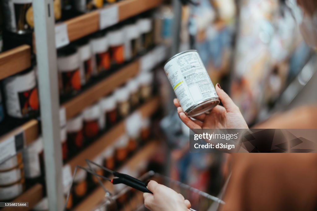 Over the shoulder view of young Asian woman carrying a shopping basket, grocery shopping in supermarket. Holding a tin can and reading the nutritional label at the back