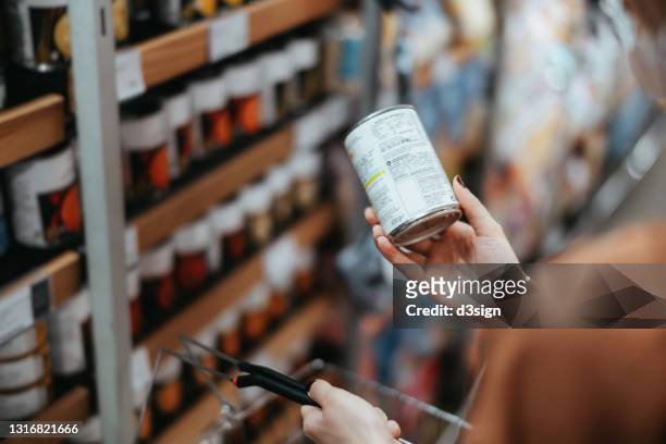 over the shoulder view of young asian woman carrying a shopping basket, grocery shopping in supermarket. holding a tin can and reading the nutritional label at the back - canned goods stock-fotos und bilder