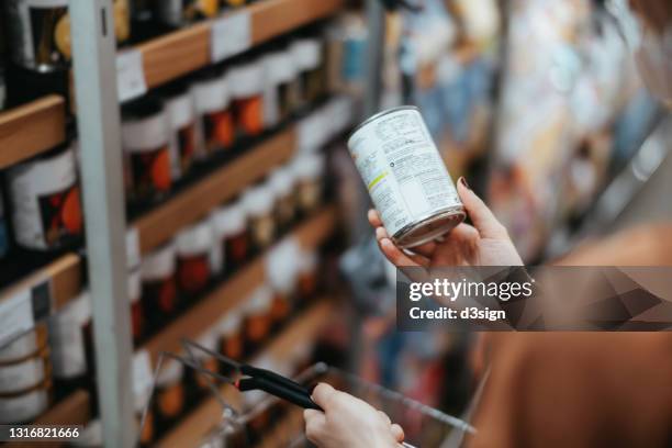 over the shoulder view of young asian woman carrying a shopping basket, grocery shopping in supermarket. holding a tin can and reading the nutritional label at the back - étiqueter photos et images de collection