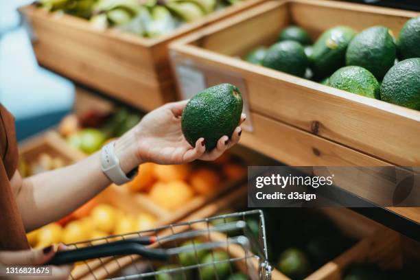 young asian woman carrying a shopping basket, grocery shopping for fresh organic fruits and vegetables along the produce aisle in supermarket, close up of her hand choosing avocados. healthy eating lifestyle - avocado stock-fotos und bilder