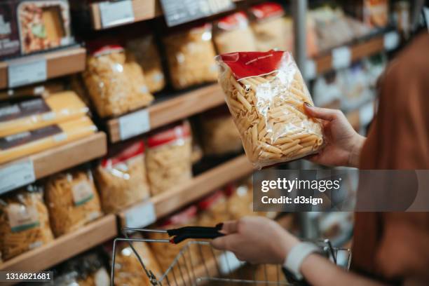 young asian woman carrying a shopping basket, grocery shopping in supermarket, close up of her hand choosing a pack of organic pasta along the aisle. healthy eating lifestyle - pasta stockfoto's en -beelden