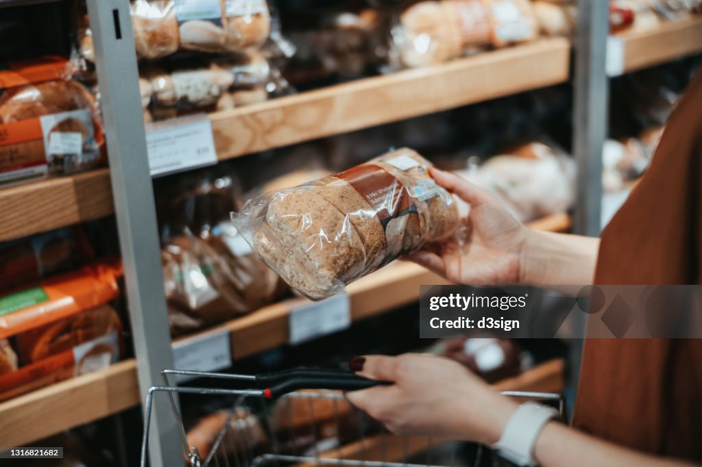 Young Asian woman carrying a shopping basket grocery shopping in a supermarket, shopping for packaged fresh wholegrain bread in the bread aisle. Healthy eating lifestyle