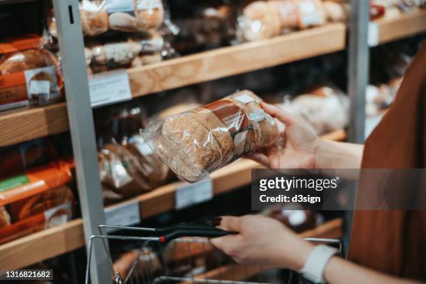 young asian woman carrying a shopping basket grocery shopping in a supermarket, shopping for packaged fresh wholegrain bread in the bread aisle. healthy eating lifestyle - bodega ストックフォトと画像