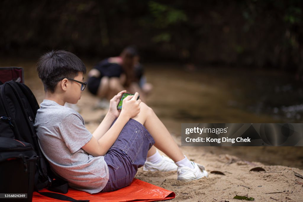 Asian Chinese teenager boy sitting alone at the riverside playing mobile games with his smart phone while his sister and mother playing in the river in the forest during weekend
