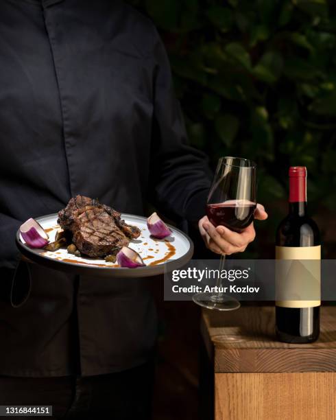 glass of red wine and plate with cooked meat in hands of unrecognizable person on black background. juicy grilled steak with onion and bottle of alcoholic beverage, close up shot - cutting red onion stock pictures, royalty-free photos & images