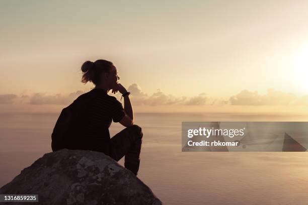 a hipster girl with a backpack sits on a high rock against the background of the sea and sky - teenager alter 個照片及圖片檔