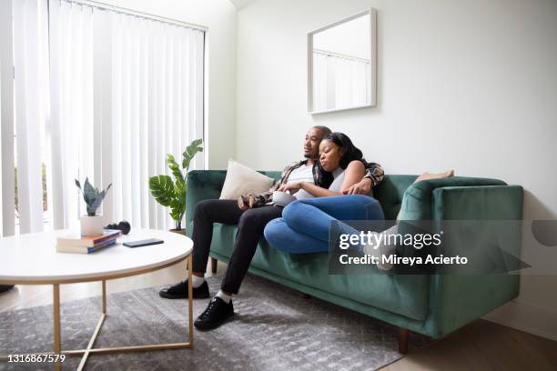a mixed race couple cuddle together on a green couch in a modern apartment, holding a remote control and a bowl of chips, while watching tv and wearing casual clothing. - young couple watching tv stock pictures, royalty-free photos & images