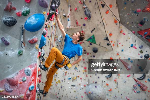 starker junger sportler skaliert kletterwand in modernem fitnessstudio - kletterwand kletterausrüstung stock-fotos und bilder