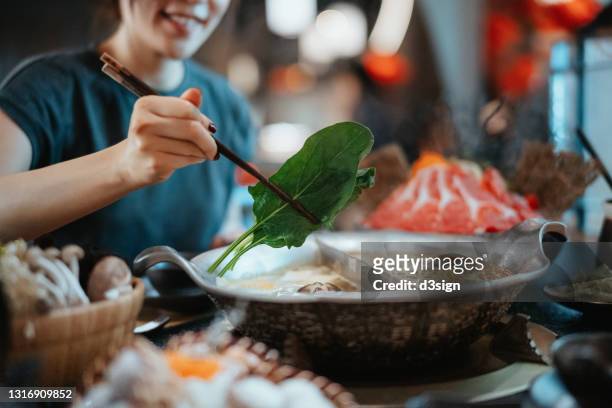 cheerful young asian woman enjoying traditional chinese hotpot with assorted fresh and scrumptious ingredients in restaurant. she is boiling vegetables into the soup. chinese culture. chinese cuisine and food. eating out lifestyle - szechuan cuisine stockfoto's en -beelden