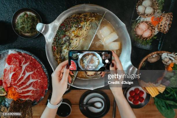 Flat lay of woman's hand taking photos of delicious traditional Chinese hotpot with assorted fresh ingredients with smartphone and sharing to social media before eating it in restaurant. Chinese cuisine. Eating out lifestyle. Camera eats first culture