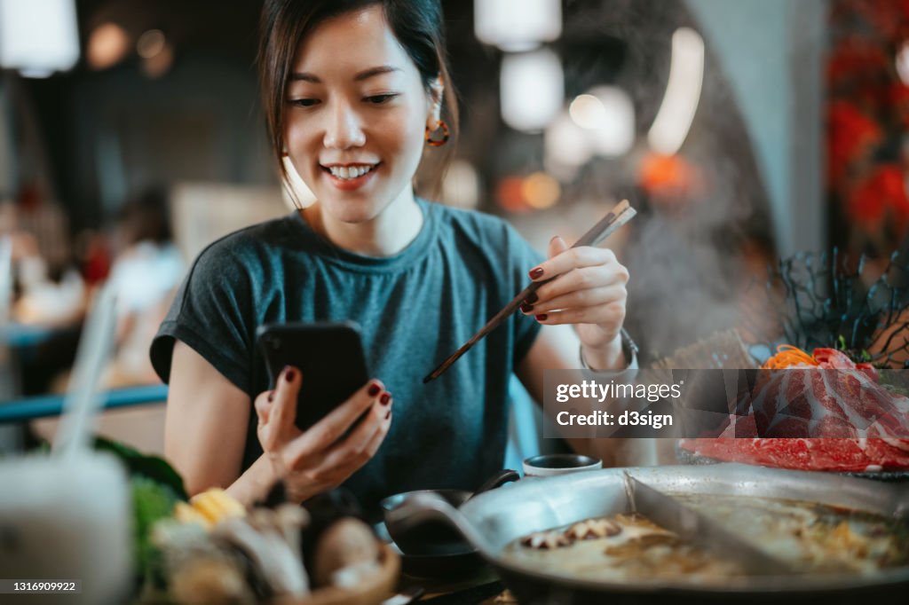 Beautiful smiling young Asian woman using smartphone while enjoying delicious traditional Chinese hotpot with assorted fresh ingredients in restaurant. Chinese cuisine and food. Eating out lifestyle. Technology in everyday life