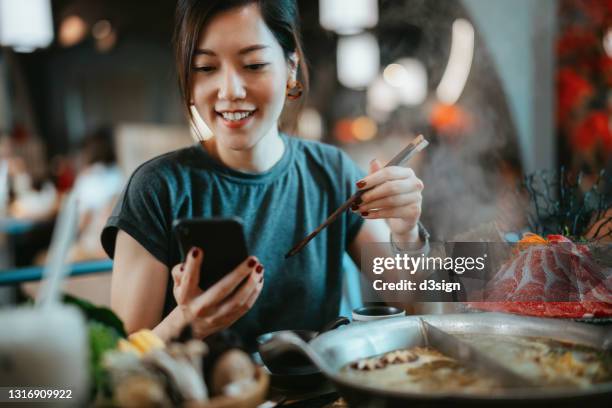 beautiful smiling young asian woman using smartphone while enjoying delicious traditional chinese hotpot with assorted fresh ingredients in restaurant. chinese cuisine and food. eating out lifestyle. technology in everyday life - hot pots stock-fotos und bilder