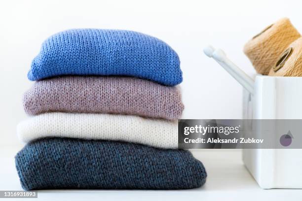 knitted things of different colors, stacked in a pile, lie on a white background. winter and autumn warm sweaters. next to the skeins and bobbins of wool yarn, knitting needles. the concept of hobby, storage, care and washing of handmade products. - wool imagens e fotografias de stock