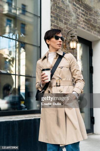 woman standing in the street holding a take away coffee in a disposable cup - trench stock photos et images de collection