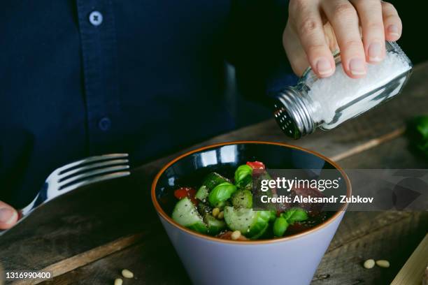 vegetarian, vegan, and raw food. a girl or woman eats a vegetable salad of tomatoes, cucumbers, nuts and seeds, green basil leaves, and salts it from a salt shaker. next to it are sliced baguette slices. the concept of healthy eating, diet. - salt shaker stockfoto's en -beelden