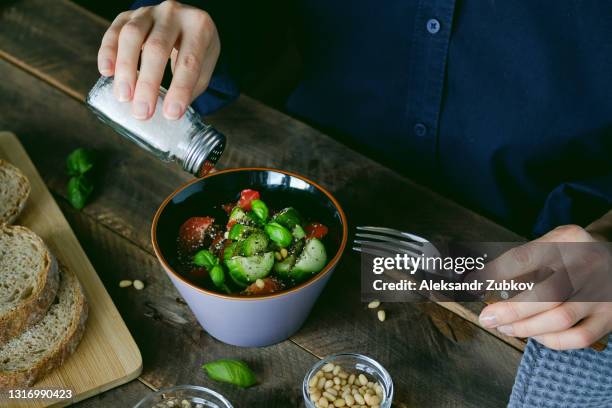 vegetarian, vegan, and raw food. a girl or woman eats a vegetable salad of tomatoes, cucumbers, nuts and seeds, green basil leaves, and salts it from a salt shaker. next to it are sliced baguette slices. the concept of healthy eating, diet. - salt shaker ストックフォトと画像