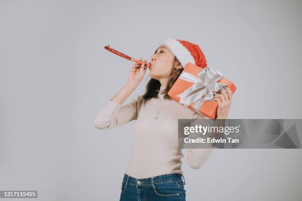 ung asiatisk kinesisk kvinna med tomtehatt blåser party hornblåsare och håller julklapp i studiogrå bakgrund - party horn blower bildbanksfoton och bilder