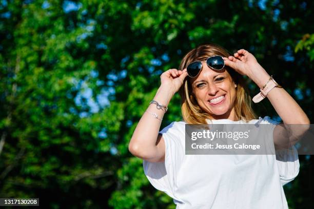 middle-aged woman, putting on her sunglasses, looking at the camera, with a park in the background. psychology and middle age concept. copyspace - positive attitude stock-fotos und bilder