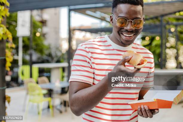 young african-american man is eating hot dog and smiling - sausage roll stock pictures, royalty-free photos & images