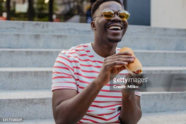 young african-american man is eating hot dog and smiling - street food stock pictures, royalty-free photos & images