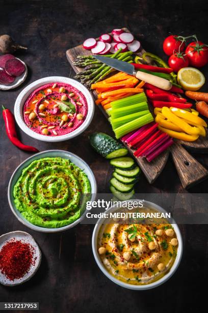 hummus three bowl of chickpeas, avocado and beetroot with cut vegetables for dip on dark wood - middle eastern food stock pictures, royalty-free photos & images