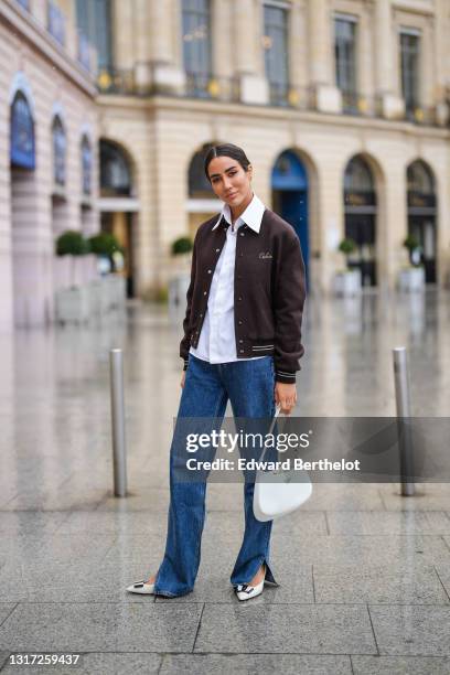 Tamara Kalinic wears a brown Celine teddy jacket, a white shirt, blue long flared slit denim jeans pants, a white Prada Cleo bag, black and white...