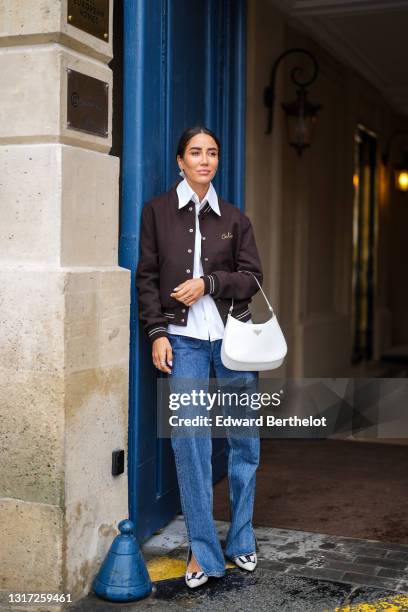 Tamara Kalinic wears a brown Celine teddy jacket, a white shirt, blue long flared slit denim jeans pants, a white Prada Cleo bag, black and white...