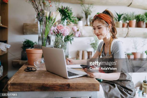 asian female florist, owner of small business flower shop, using smartphone while working on laptop against flowers and plants. checking stocks, taking customers orders, selling products online. daily routine of running a small business with technology - asian females on a phone imagens e fotografias de stock