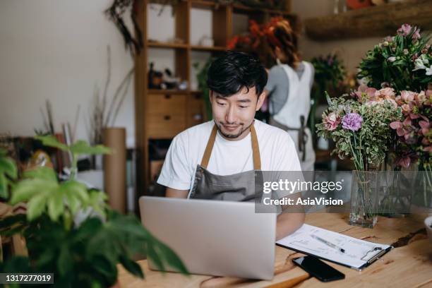 young asian couple, owners of small business flower shop, working on laptop over counter next to flowers and plants. checking stocks, taking customer orders, selling products online. daily routine of running a small business with technology - asia lady selling flower stock pictures, royalty-free photos & images