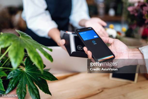 close up of young asian man shopping at the flower shop. he is paying with his smartphone, scan and pay a bill on a card machine making a quick and easy contactless payment. nfc technology, tap and go concept - contactless payment stock pictures, royalty-free photos & images