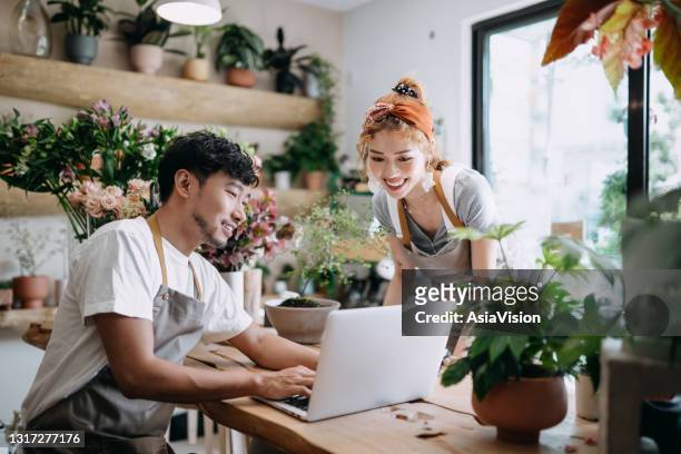 smiling young asian couple, the owners of small business flower shop, discussing over laptop on counter against flowers and plants. start-up business, business partnership and teamwork. working together for successful business - small business stock pictures, royalty-free photos & images