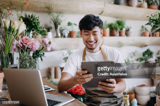 asian male florist, owner of small business flower shop, using digital tablet while working on laptop against flowers and plants. checking stocks, taking customer orders, selling products online. daily routine of running a small business with technology - new business stock pictures, royalty-free photos & images