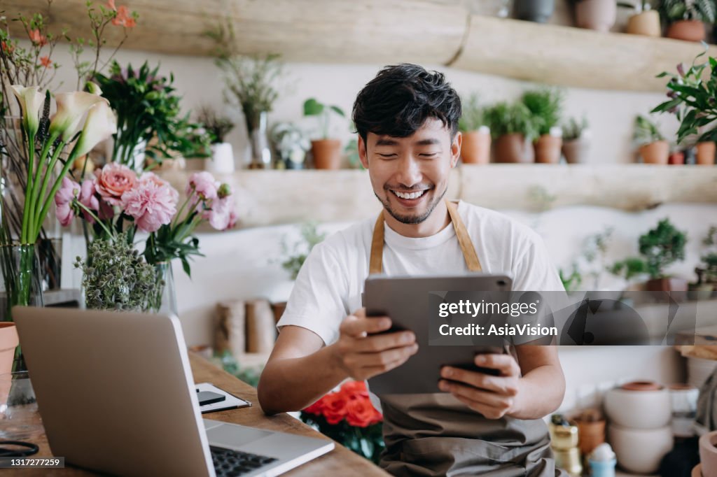 Asian male florist, owner of small business flower shop, using digital tablet while working on laptop against flowers and plants. Checking stocks, taking customer orders, selling products online. Daily routine of running a small business with technology