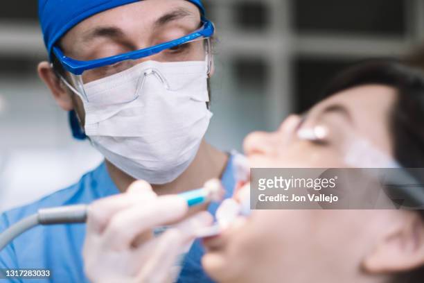 el dentista está tratando la boca de una mujer joven con gafas de plástico contra salpicaduras y un separador dental. enfoque selectivo sobre el dentista que lleva puestas sus gafas antisalpicaduras, su mascarilla y su bata azul. - mascarilla stockfoto's en -beelden