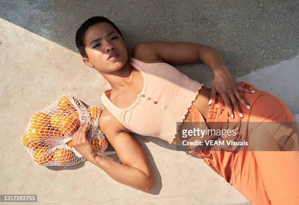 short hair woman lying on the floor dressed in orange tones and carrying a bag full of oranges. - fashion orange colour stock pictures, royalty-free photos & images