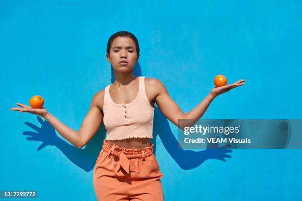 young short hair woman dressed with a orange crop top and orange pants holding one orange with each hand in front of a blue wall. - hands in her pants fotografías e imágenes de stock