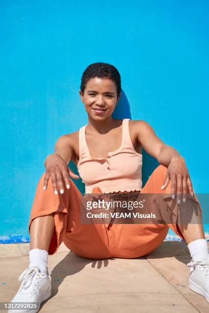 stylish short hair woman dressed in orange crop top and pants and white sport shoes sitting on the floor against a blue wall. - orange pants stockfoto's en -beelden