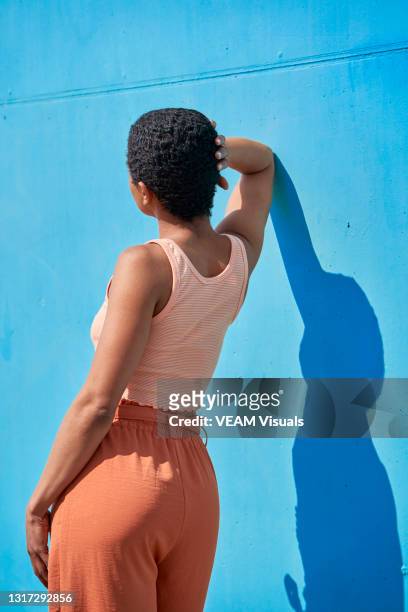 view of the back of a short-hair woman dressed in orange colors leaning with her elbow on a blue painted wall. - navel orange stockfoto's en -beelden