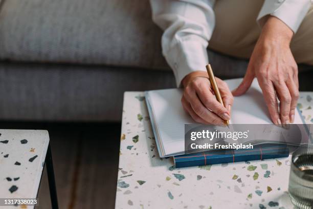 anonymous senior woman sitting in her living room and taking notes on a coffee table - 50s woman writing at table stock pictures, royalty-free photos & images