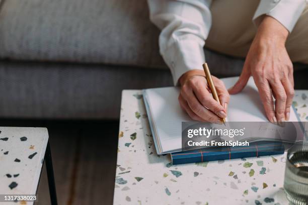 anonymous senior woman sitting in her living room and taking notes on a coffee table - 50s woman writing at table imagens e fotografias de stock