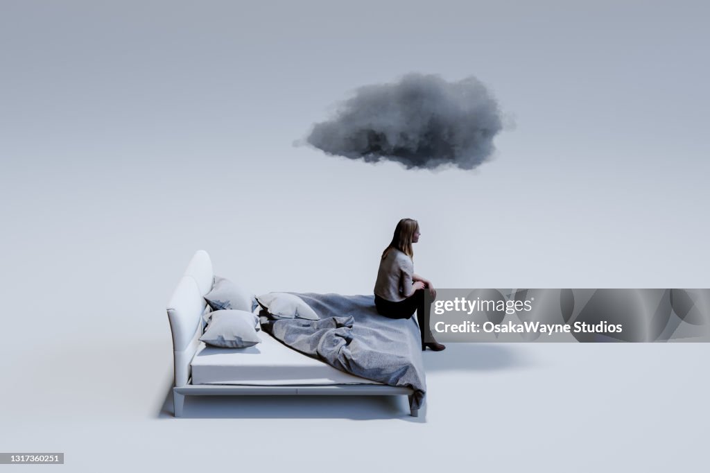 Sad mid adult woman sitting on corner of double bed with dark cloud above head.