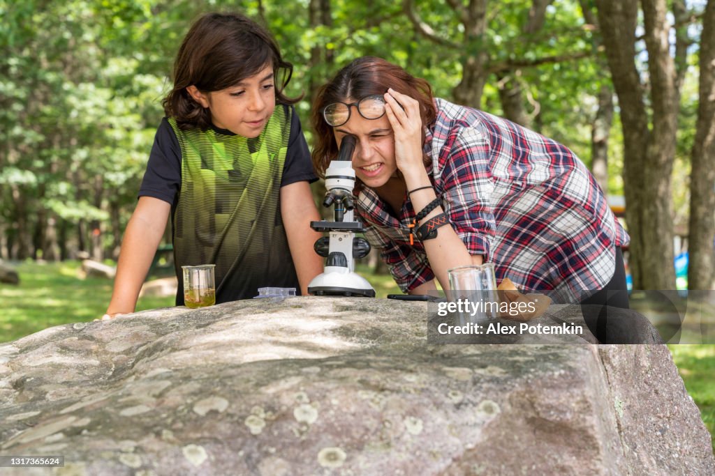 Teenage girl, 17-years-old sister teaching her 9-years-old younger brother how to working with a microscope and learning nature outdoors at the lakeshore in the sunny summer day.