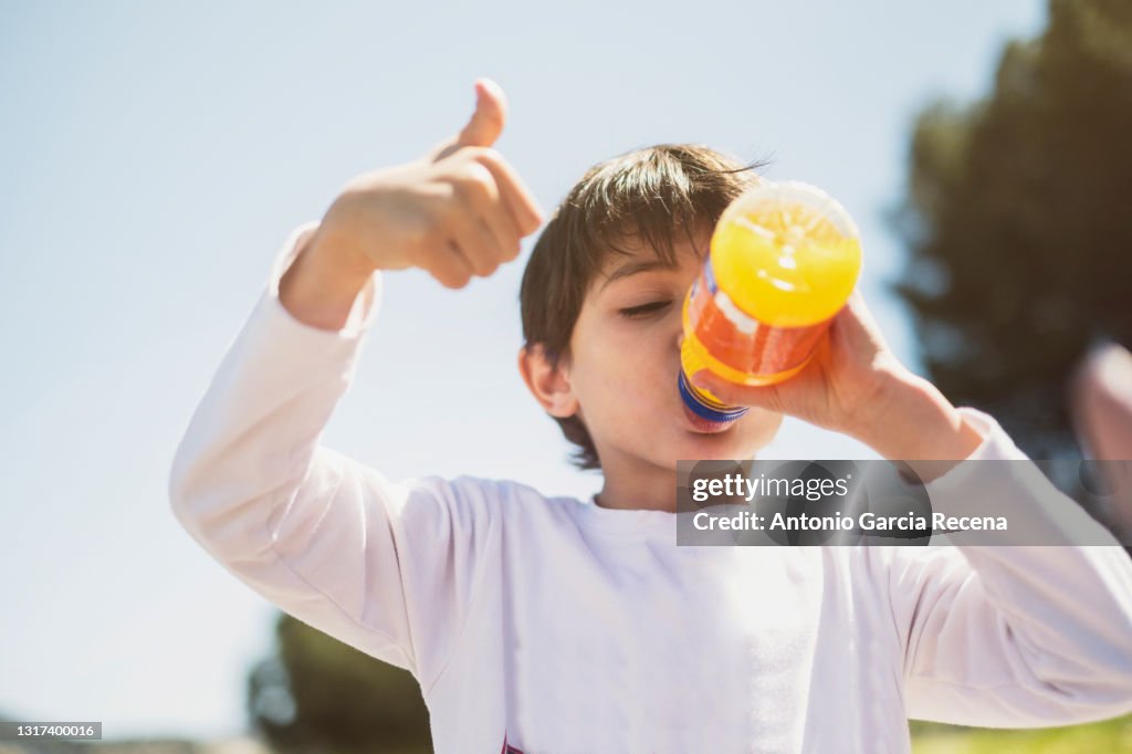A happy child makes the ok symbol with the sky in the background while drinking industrial orange juice